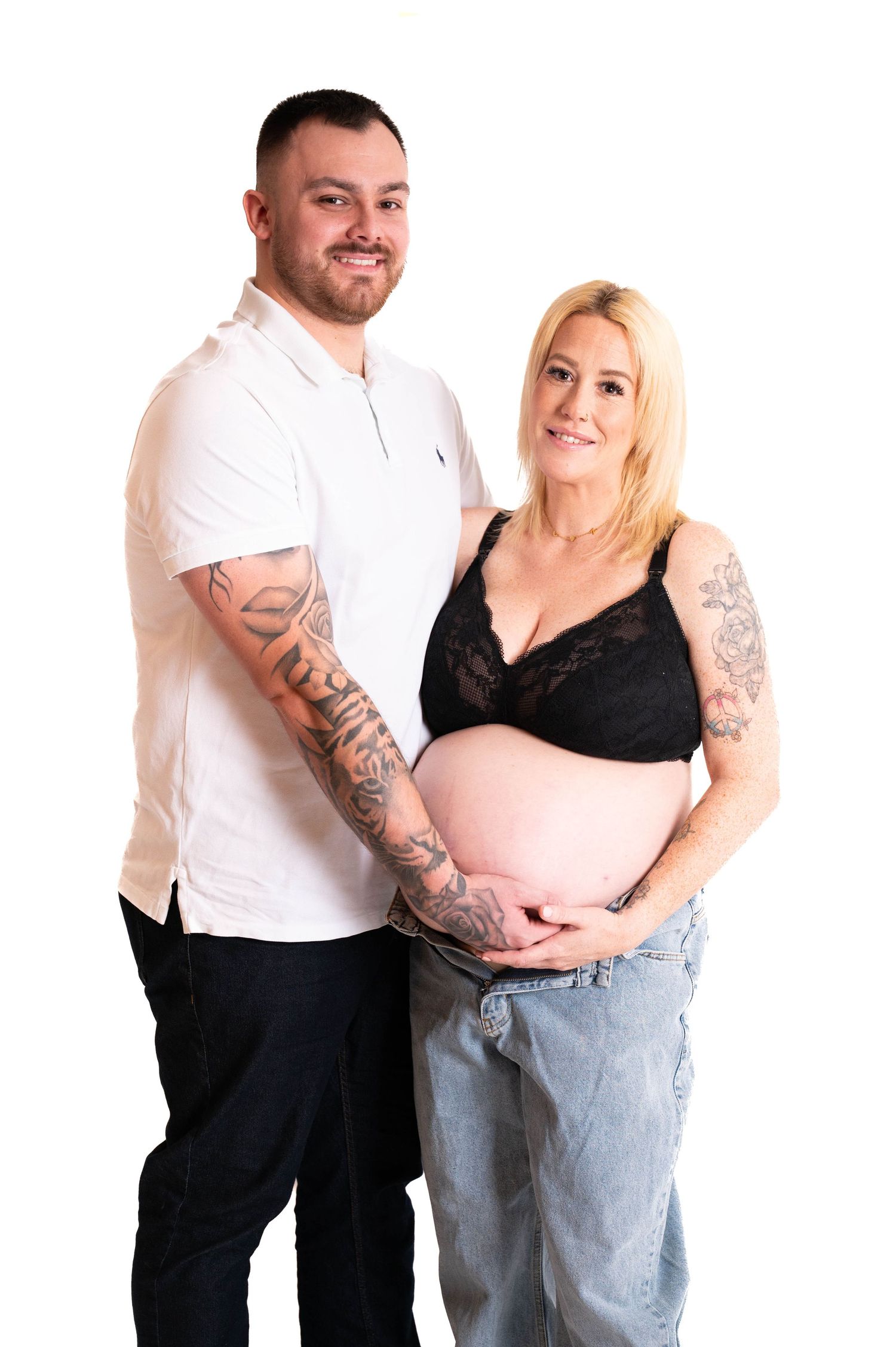 Expecting couple in casual attire pose together for maternity photo against white background.