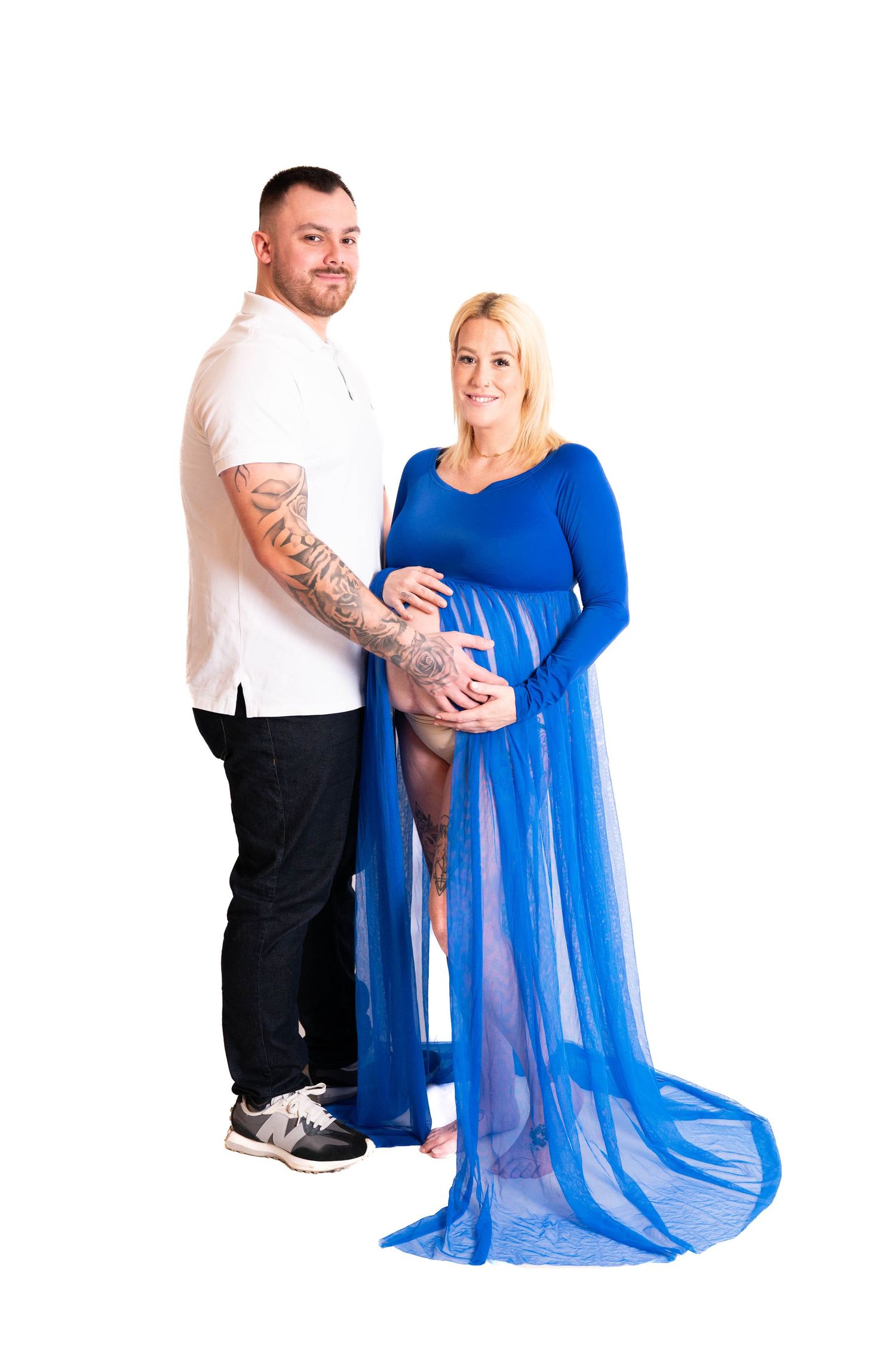 Couple poses for maternity photo with woman wearing flowing blue dress against white backdrop.