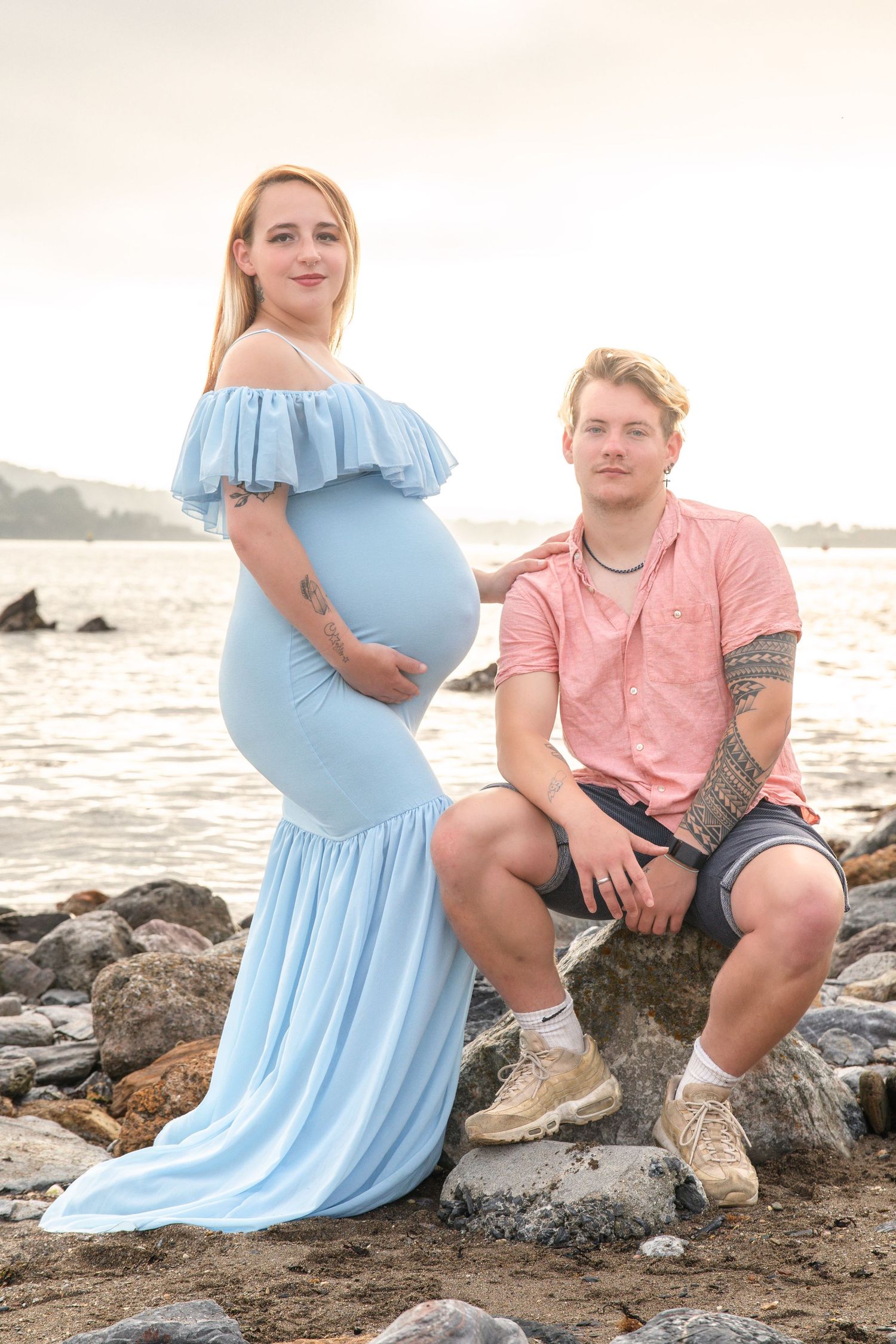 A couple poses on rocky beach at sunset, with the woman wearing a flowing light blue maternity dress.
