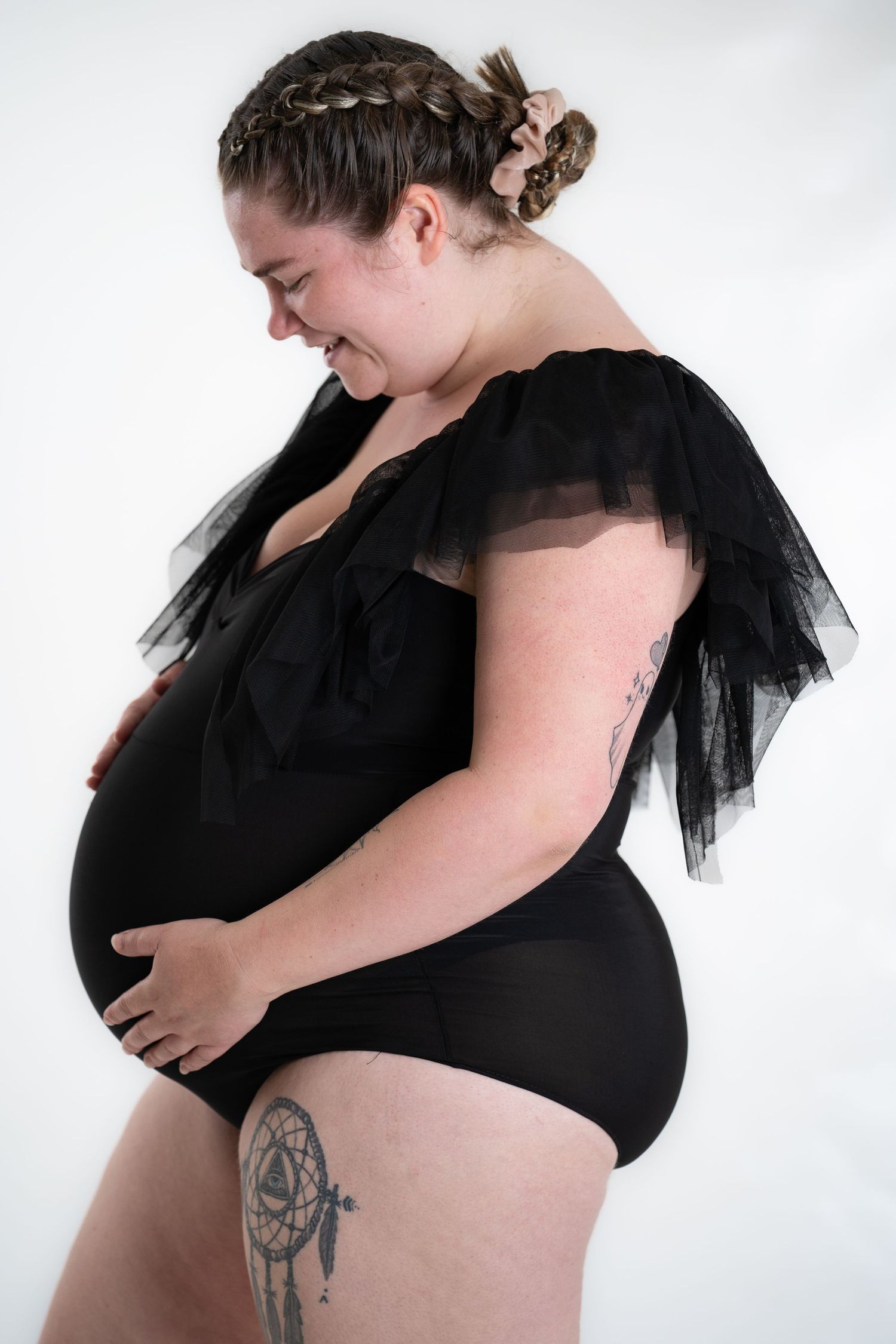 Maternity portrait in black tulle bodysuit showing baby bump against white background.
