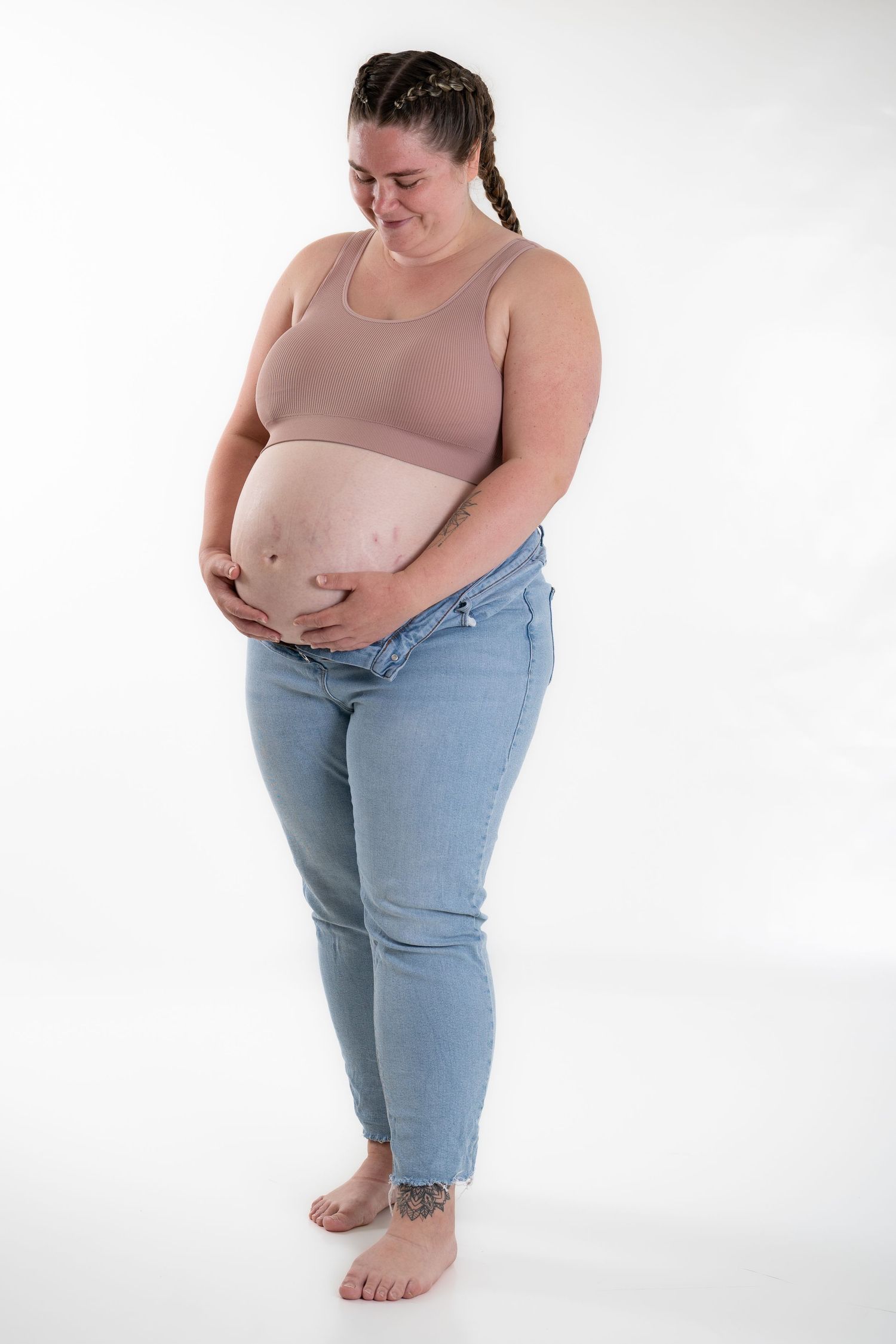 Pregnant person in light tank top and jeans poses against white background showing baby bump.