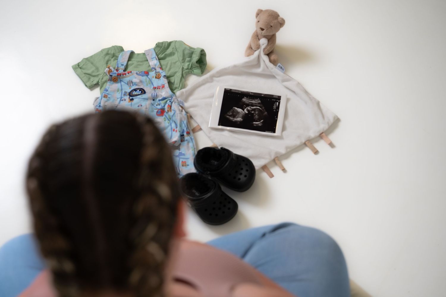 Flatlay of baby items including onesie, ultrasound photo, and teddy bear on white background.