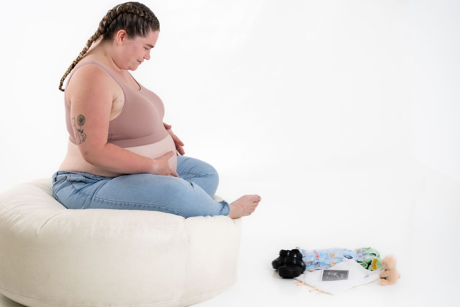 Expectant person sits on white pouf with baby items displayed nearby.
