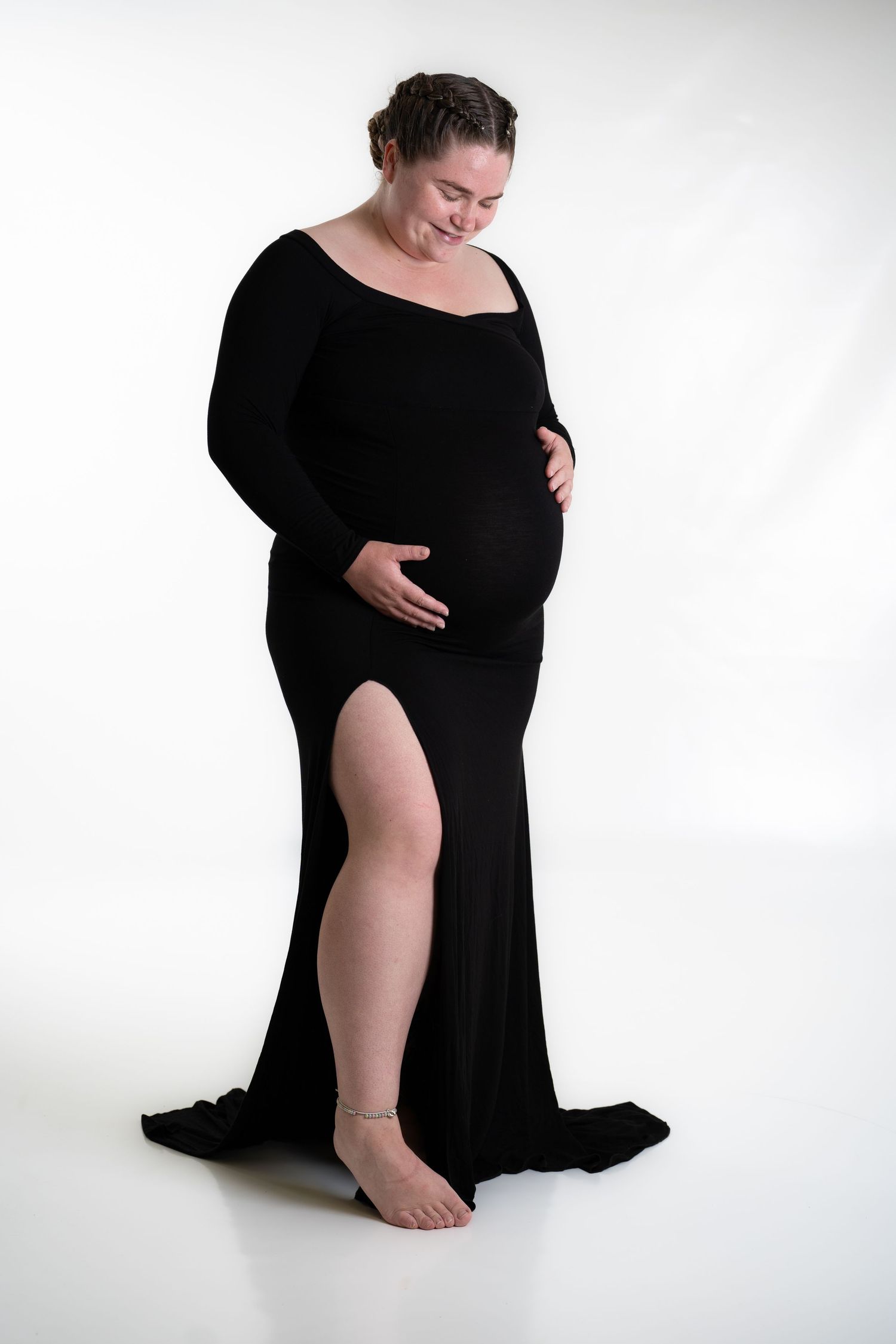 Maternity photoshoot featuring elegant black dress with thigh slit against white backdrop.
