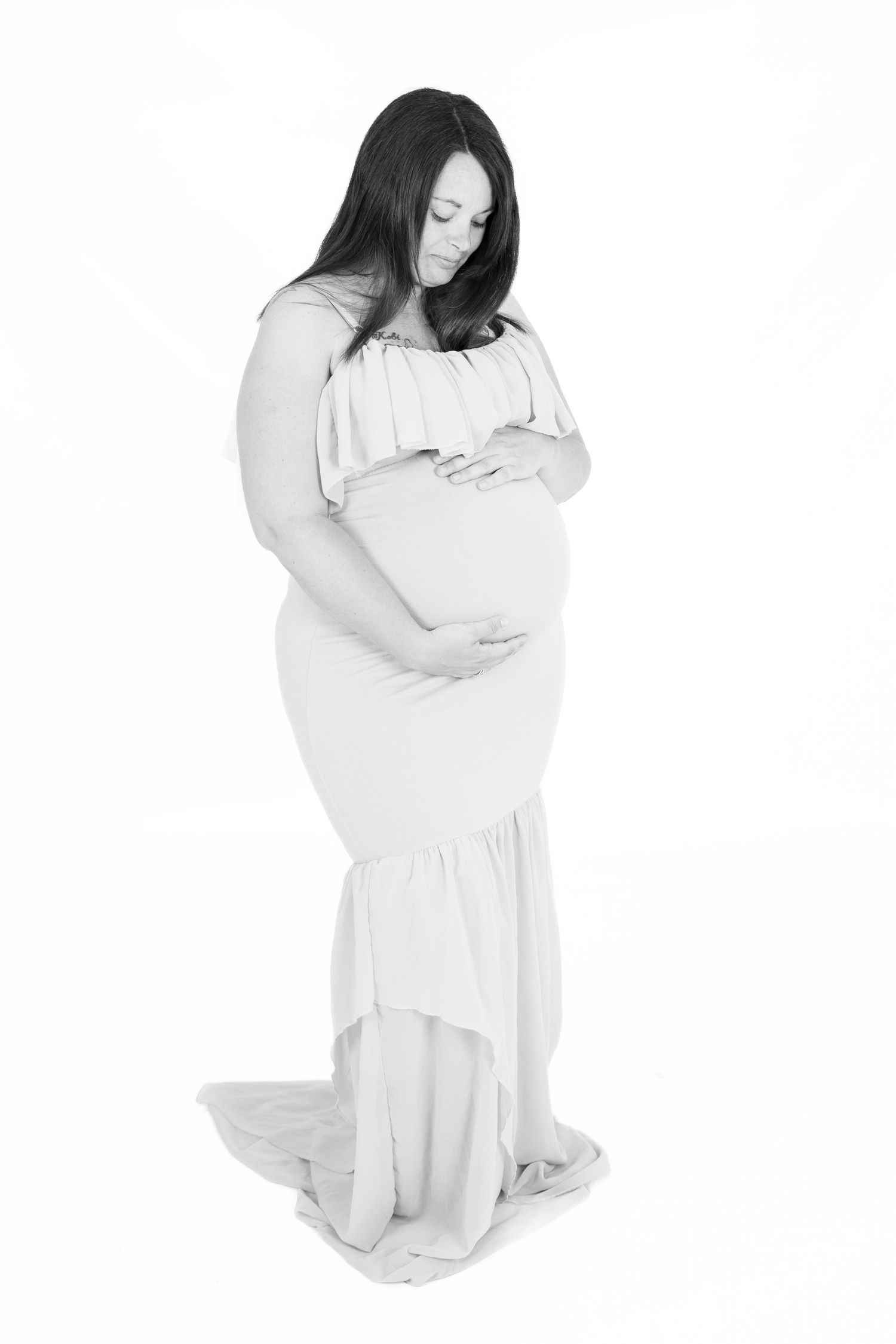 Black and white portrait of an expectant mother in a flowing white dress gently cradling her baby bump.