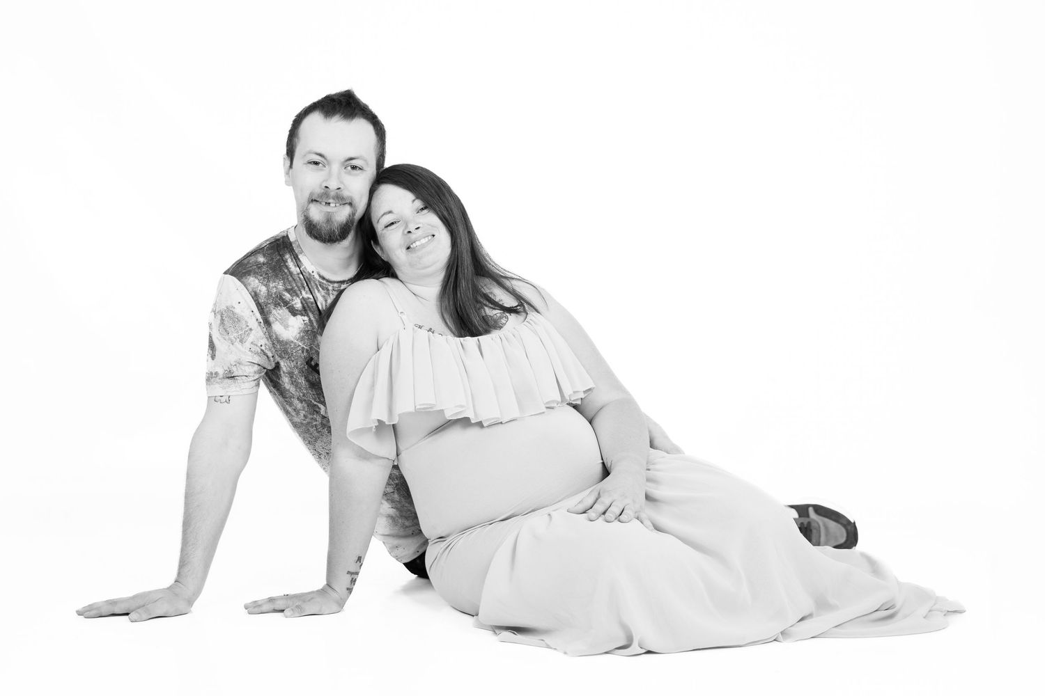 Black and white studio photo of a couple sitting together on the floor in light colored clothing.
