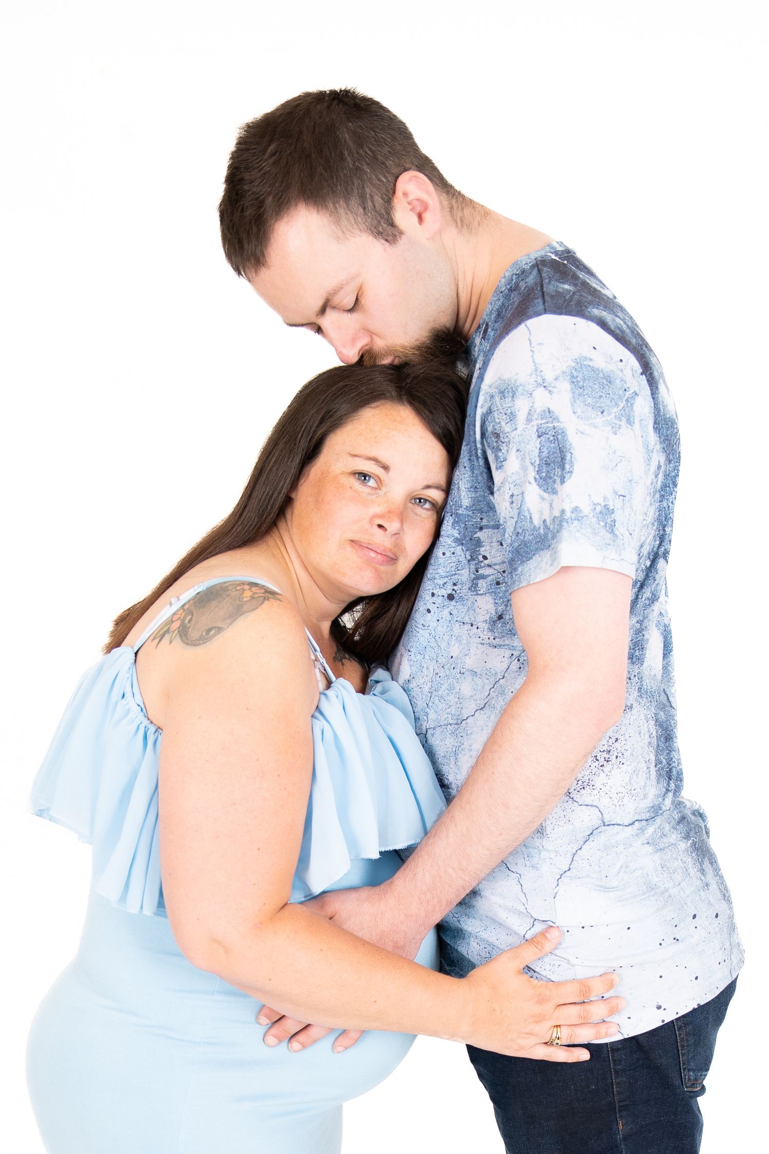 A pregnant woman in a blue top embraces with her partner who wears a patterned shirt against a white background.