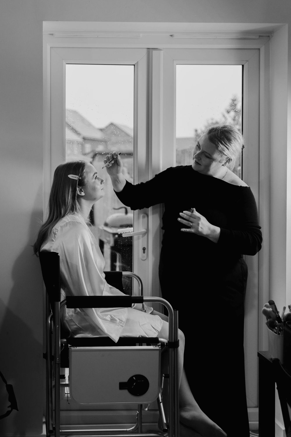 A stylist works on a client's makeup by a window in a monochrome setting.