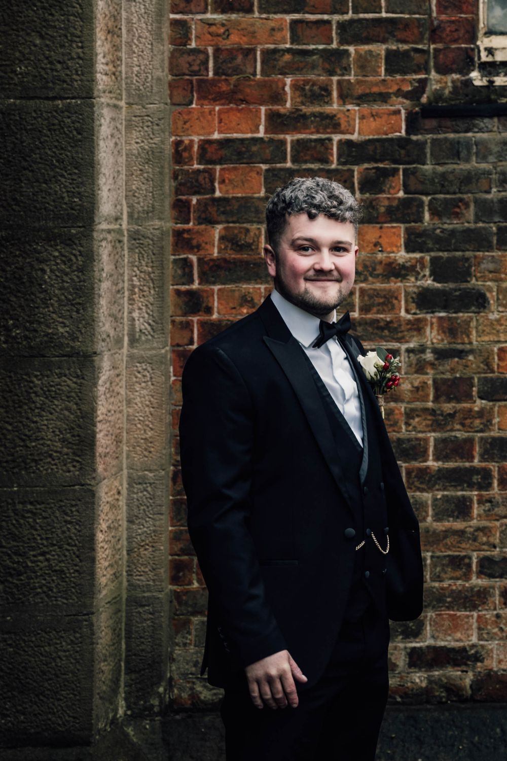 Groom in classic black tuxedo poses against rustic brick wall.