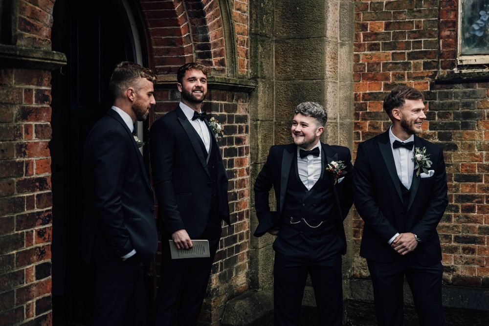 Groomsmen in black tuxedos gathered against brick wall exterior.