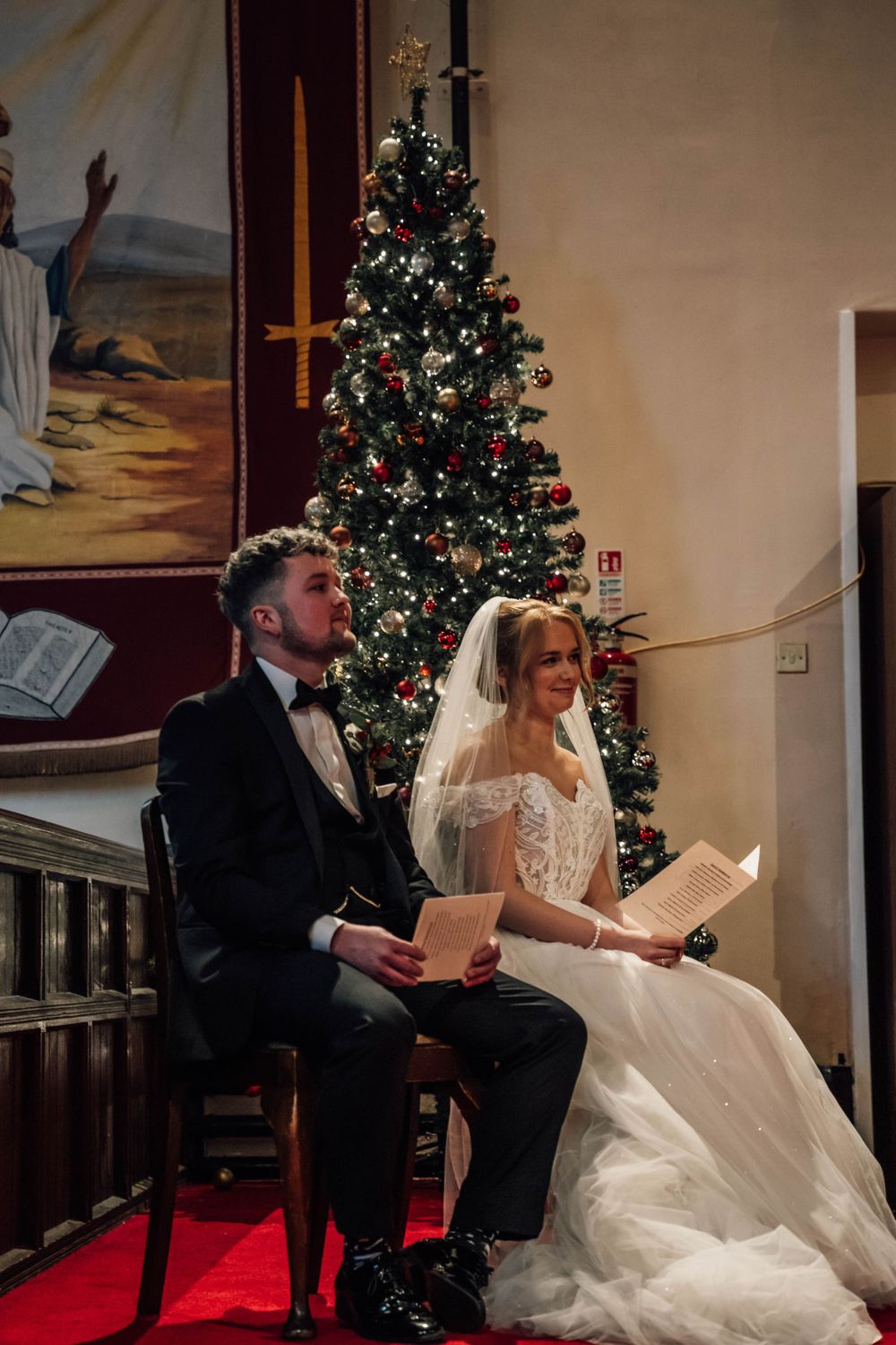 A couple sits on red chairs during their winter wedding ceremony with a Christmas tree behind them.