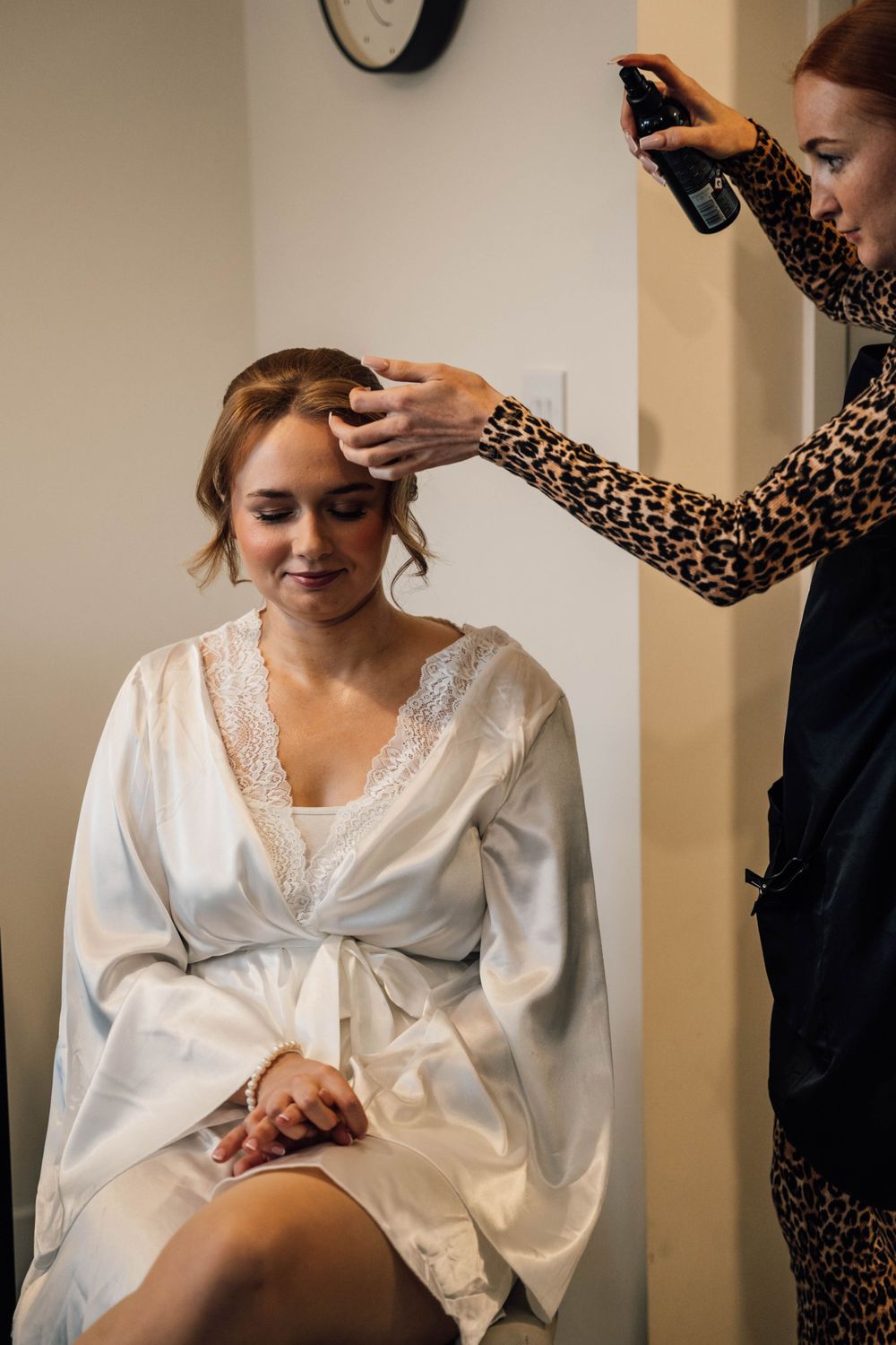 Bride in white silk robe getting hair and makeup done during wedding preparations.