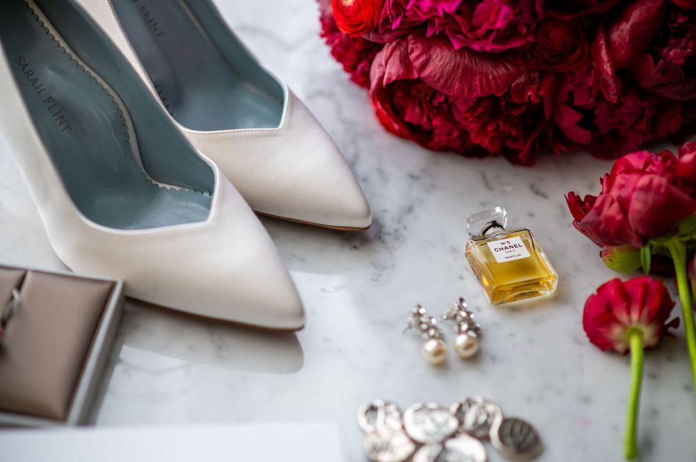 White bridal shoes with perfume bottle, earrings and red flowers on marble surface.