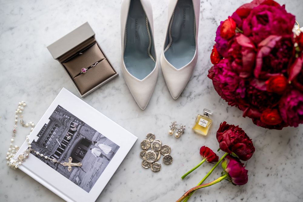 Wedding details flatlay with ivory heels, red bouquet, jewelry and photo album on marble.