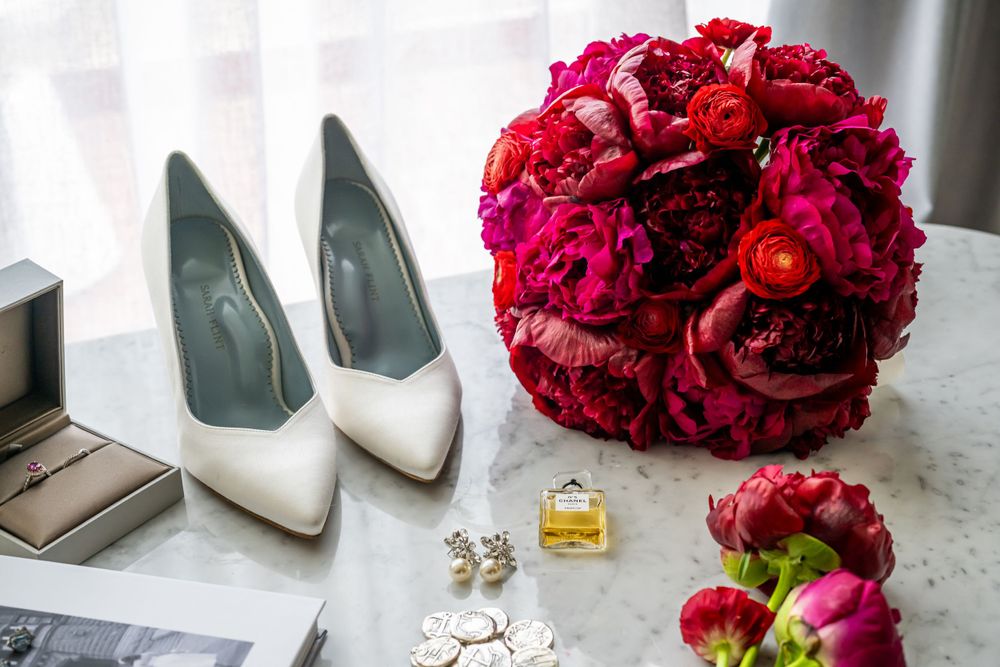 Wedding styling with white heels, red rose bouquet and accessories on marble surface.