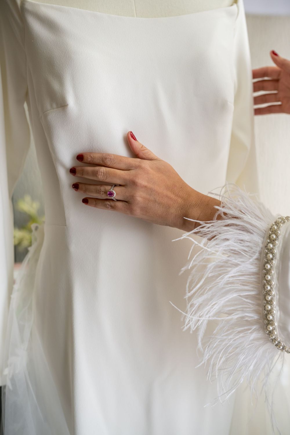 Close-up of hand with ring touching white wedding dress with feather detail.
