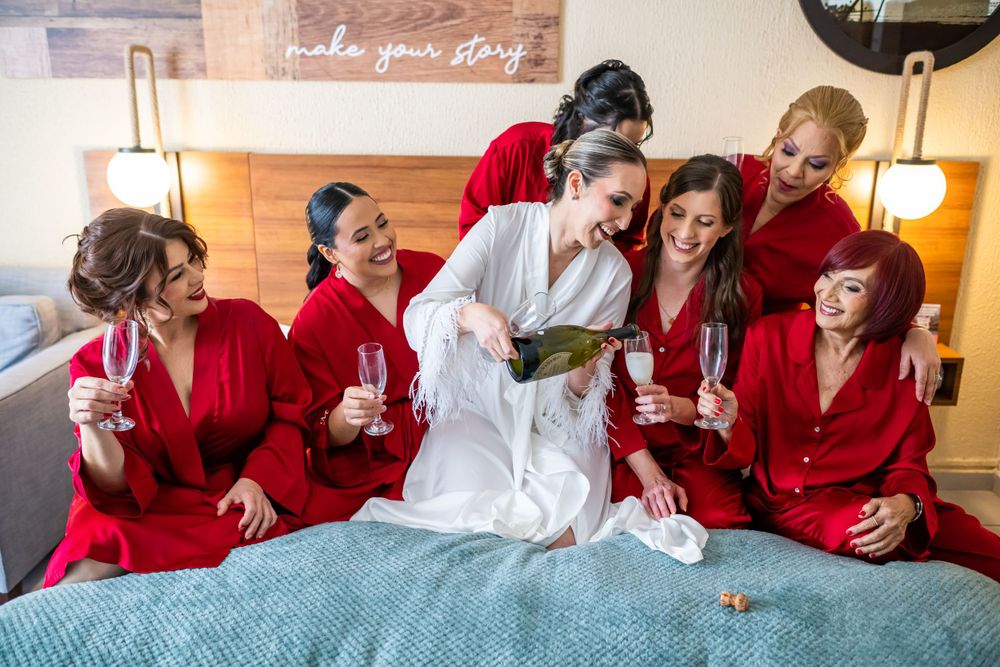 Bride in white robe surrounded by bridesmaids in red robes sharing champagne and laughter on wedding morning.