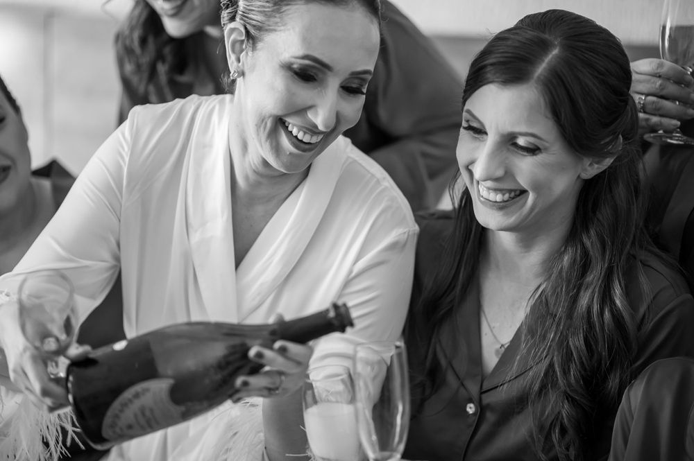 Black and white photo of women sharing champagne and joyful moments during wedding preparations.