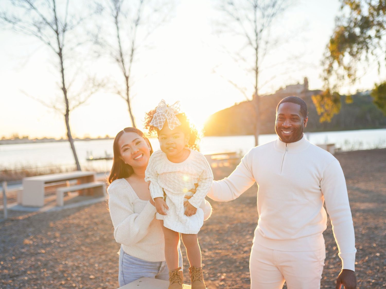 A joyful family moment captured in sunlight as parents lift their child while playing near a lake.
