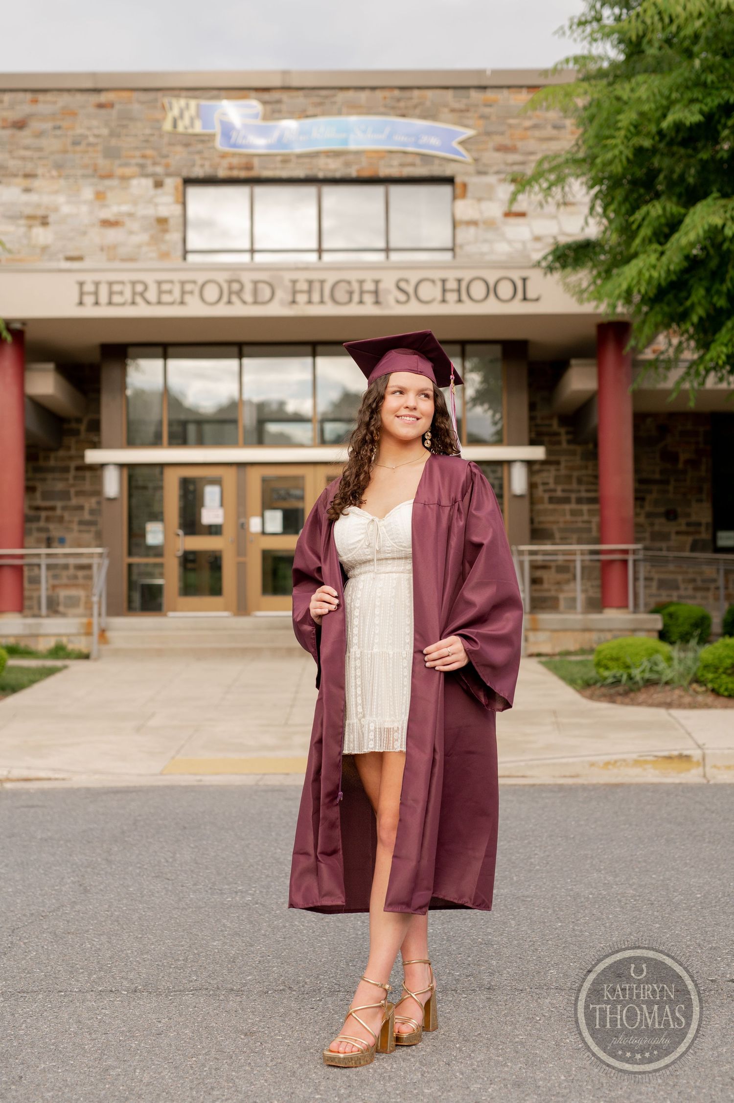 Graduate stands proudly in burgundy graduation attire outside Hereford High School entrance.