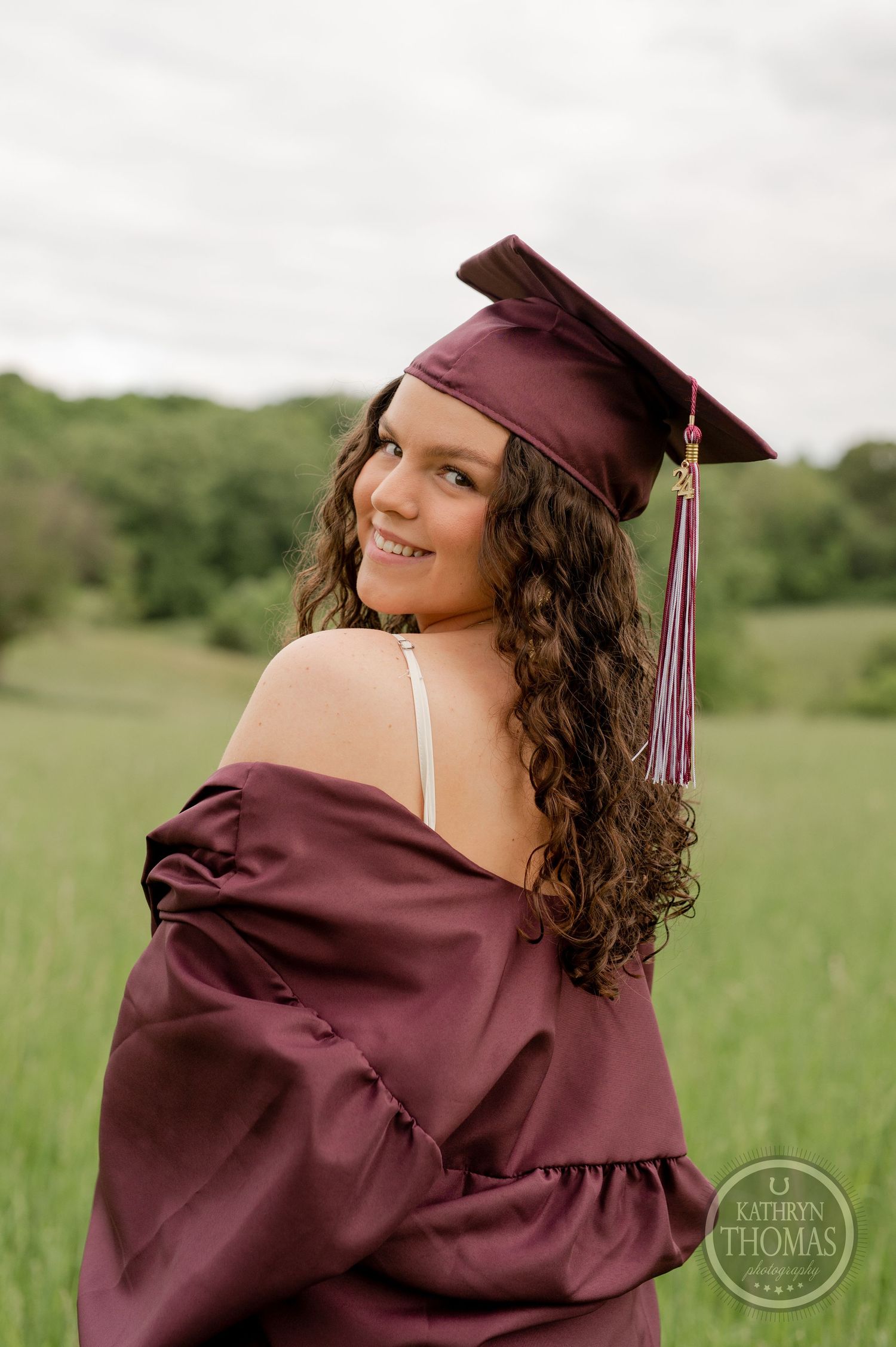 Graduate in maroon cap and gown poses over shoulder in a grassy field at sunset.