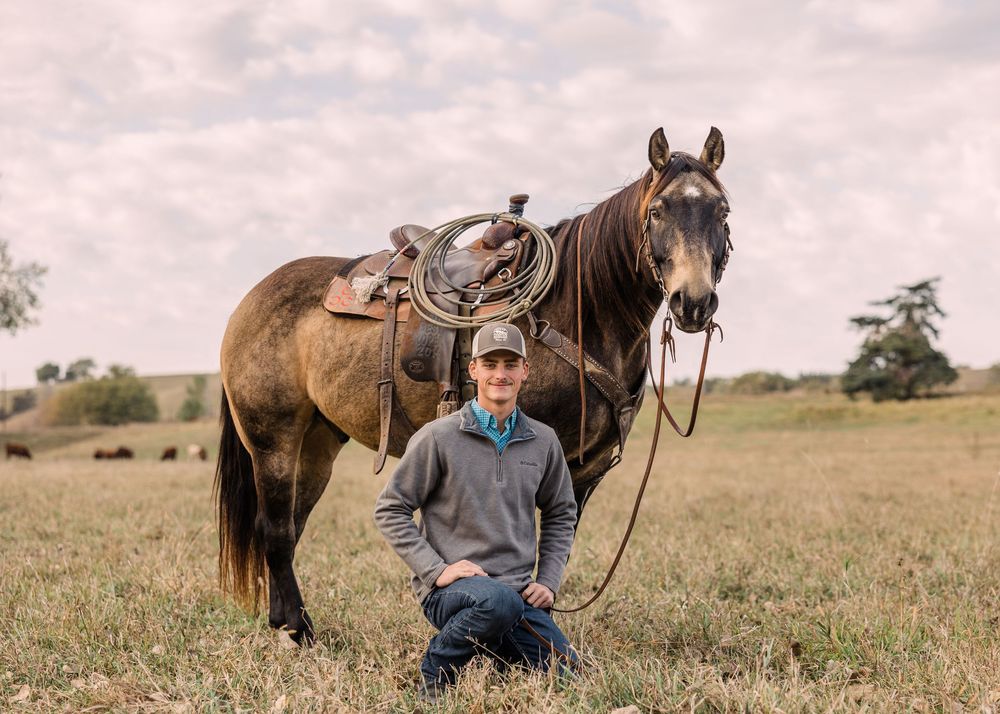 Young man in a cap kneels in a grassy pasture beside a saddled buckskin horse under a soft cloudy sky.