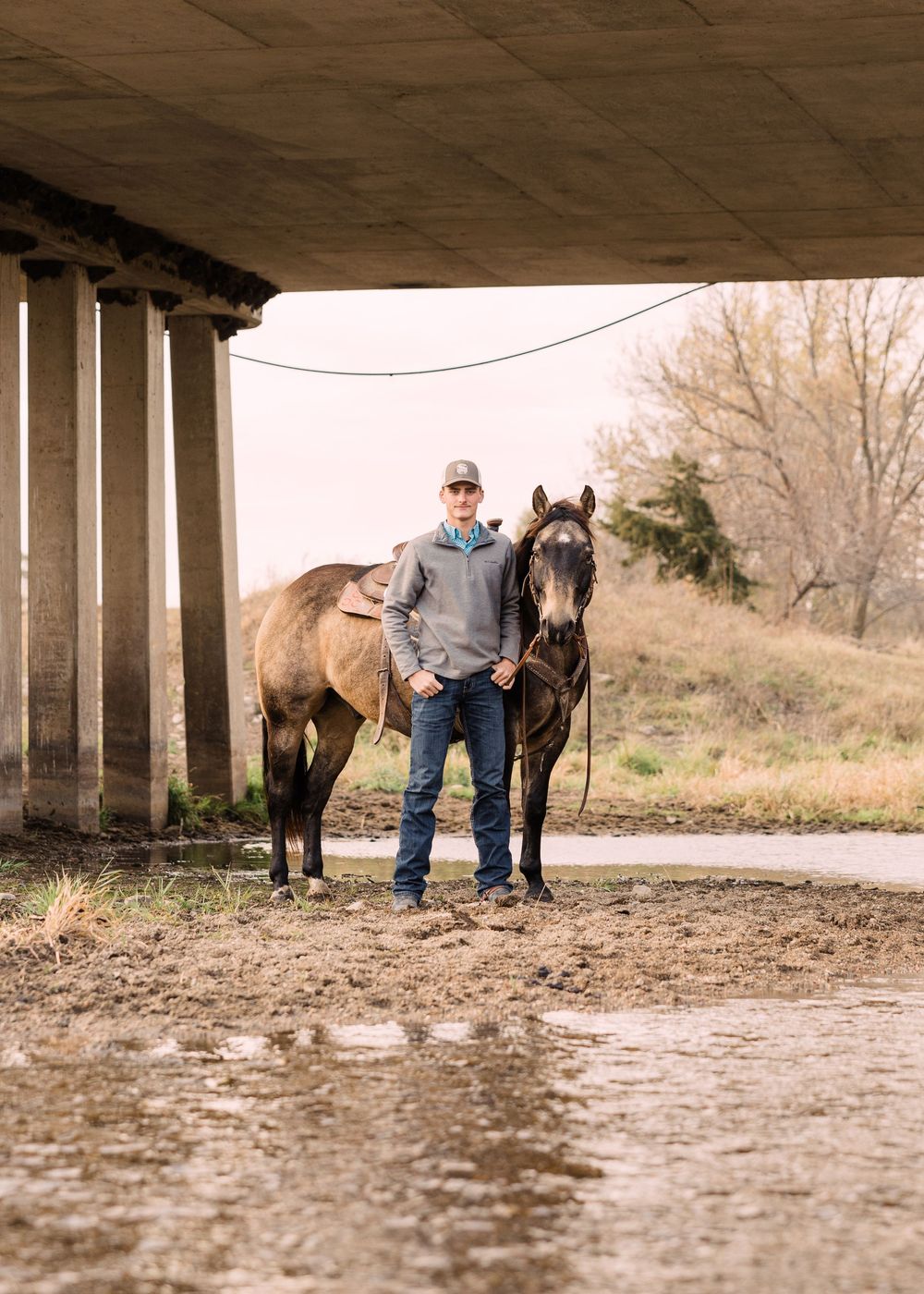Man in a gray hoodie and ball cap stands with his saddled buckskin horse beneath a concrete bridge beside a muddy creek.