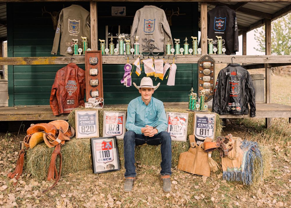 Cowboy in a blue shirt and hat sits on hay bales surrounded by rodeo trophies, buckles, saddles and show jackets.