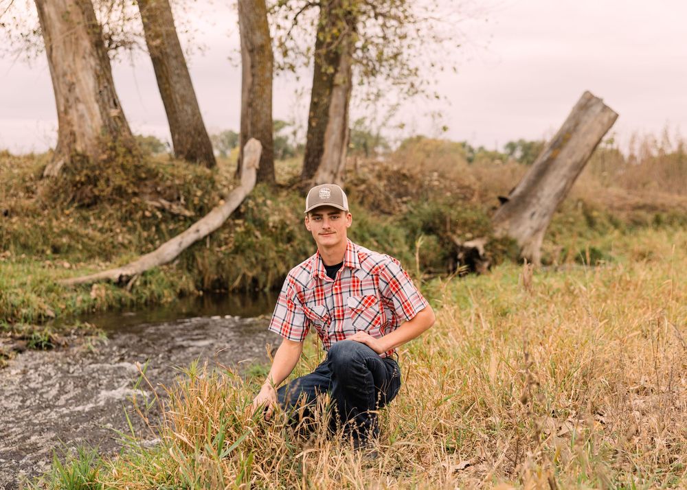 Young man in a plaid shirt and cap crouches on a grassy bank beside a small rocky creek surrounded by weathered trees.
