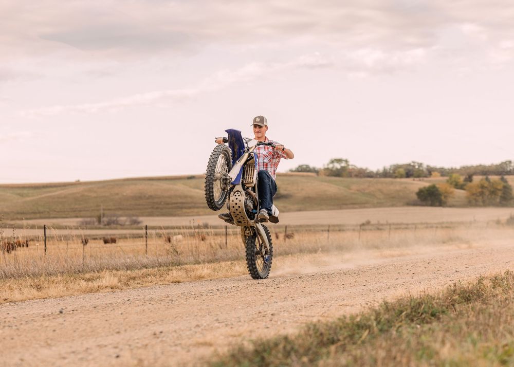 Rider in a cap pops a wheelie on a dirt bike along a dusty gravel road with rolling prairie hills under a soft pink sky.