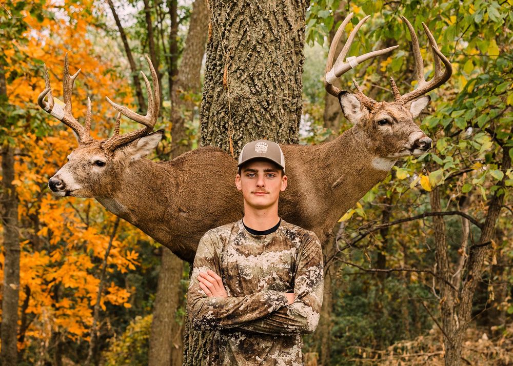 Young hunter in camouflage and a ball cap stands with arms crossed in front of a large whitetail buck mount in autumn woods.