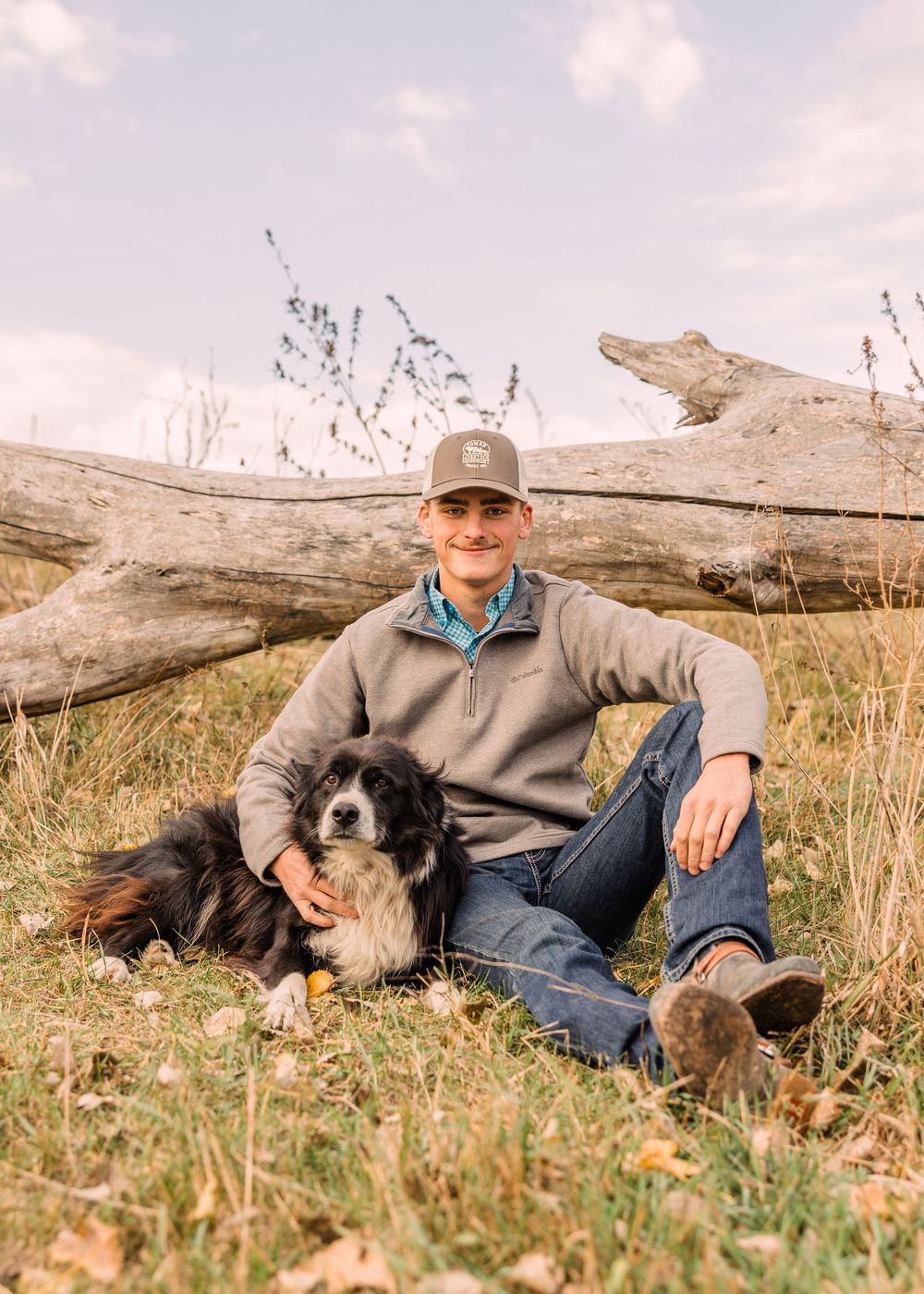 Young man in a baseball cap sits in dry grass with his black and white border collie beside a large fallen log.