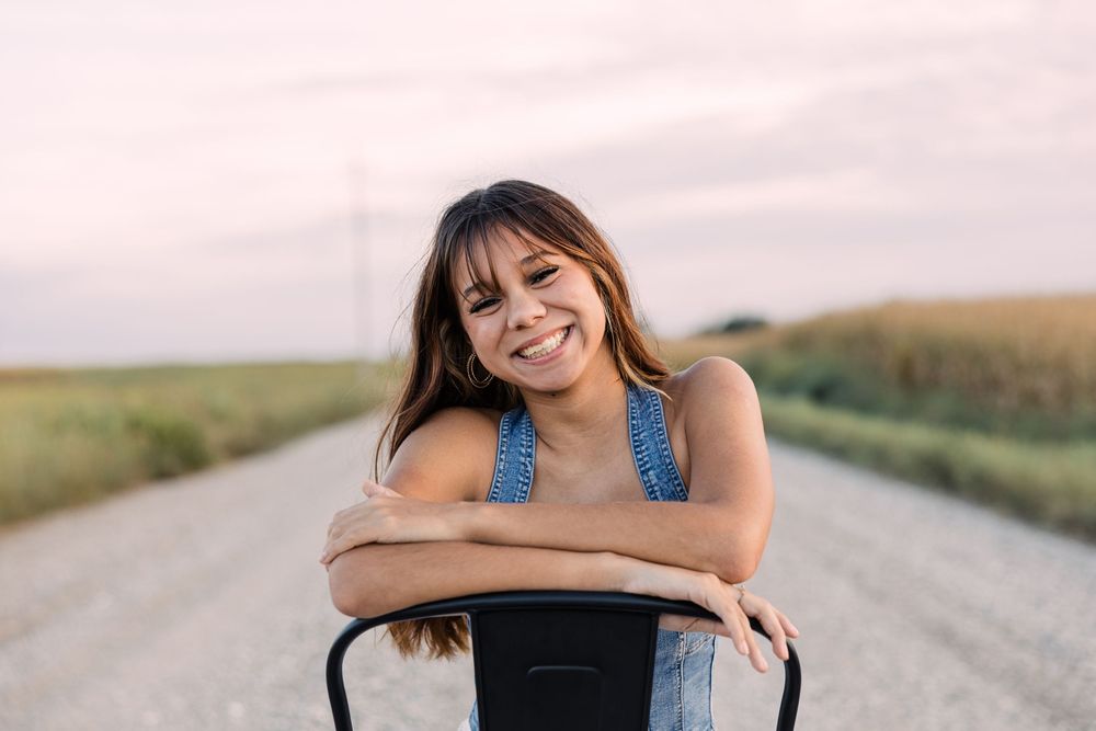 Smiling young woman in a denim top leans on the back of a black metal chair on a gravel road beside golden crop fields.