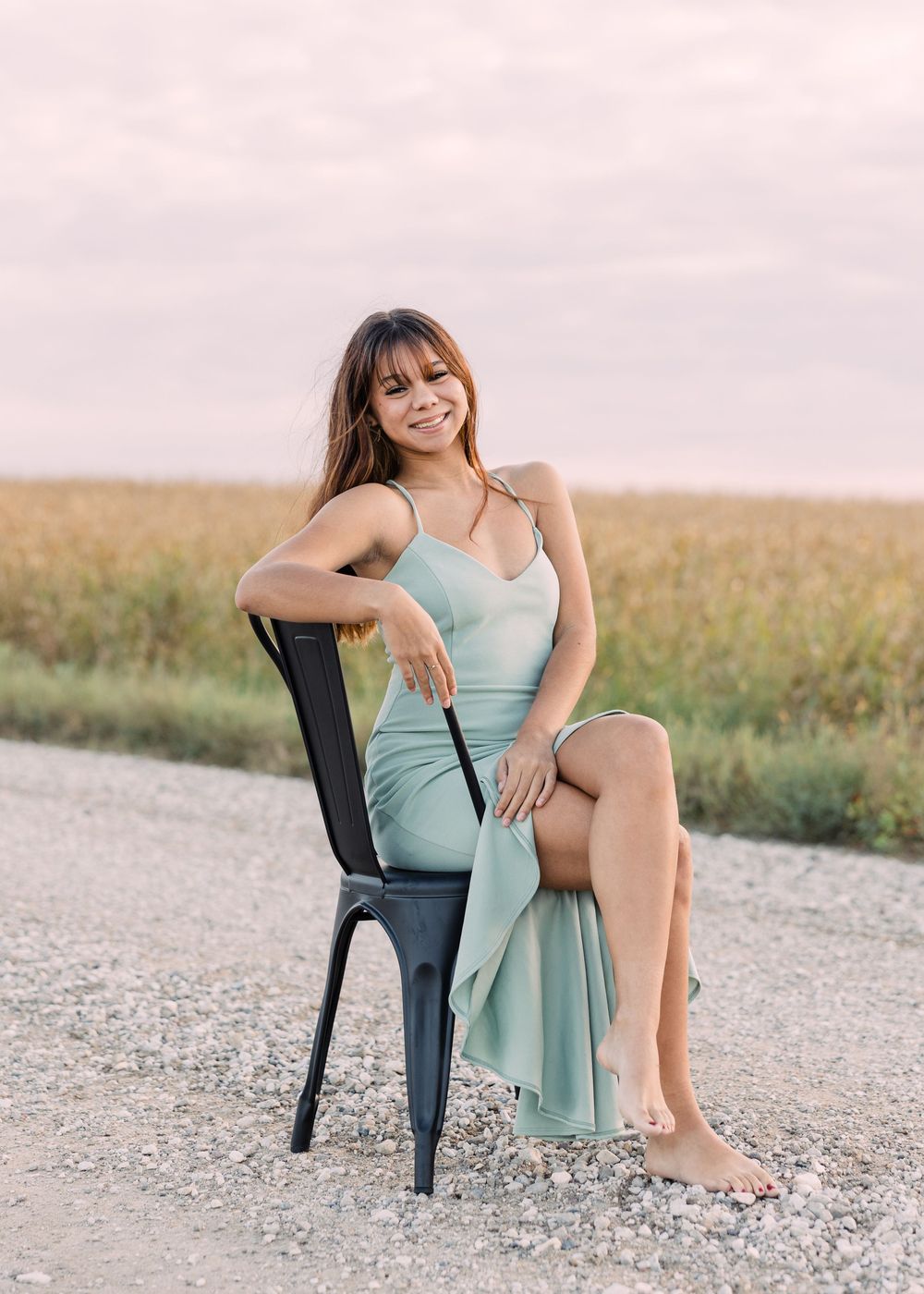 Young woman in a flowing sage green dress sits barefoot on a black metal chair on a gravel road in front of a golden field.