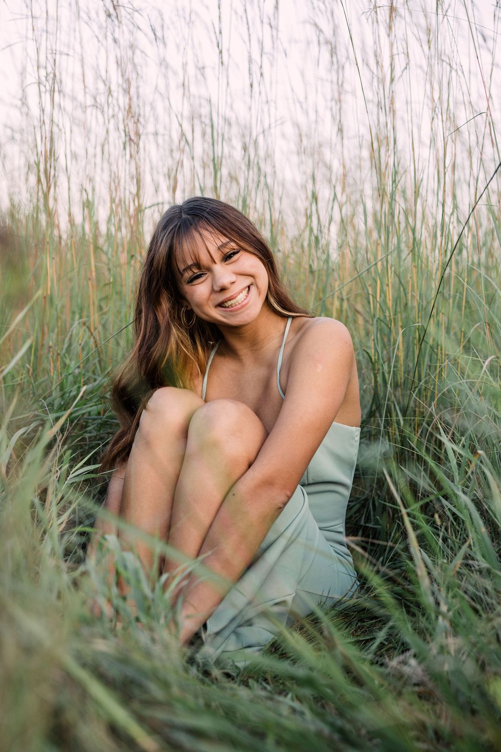 Smiling young woman with long brown hair sits hugging her knees in a field of tall green grass.