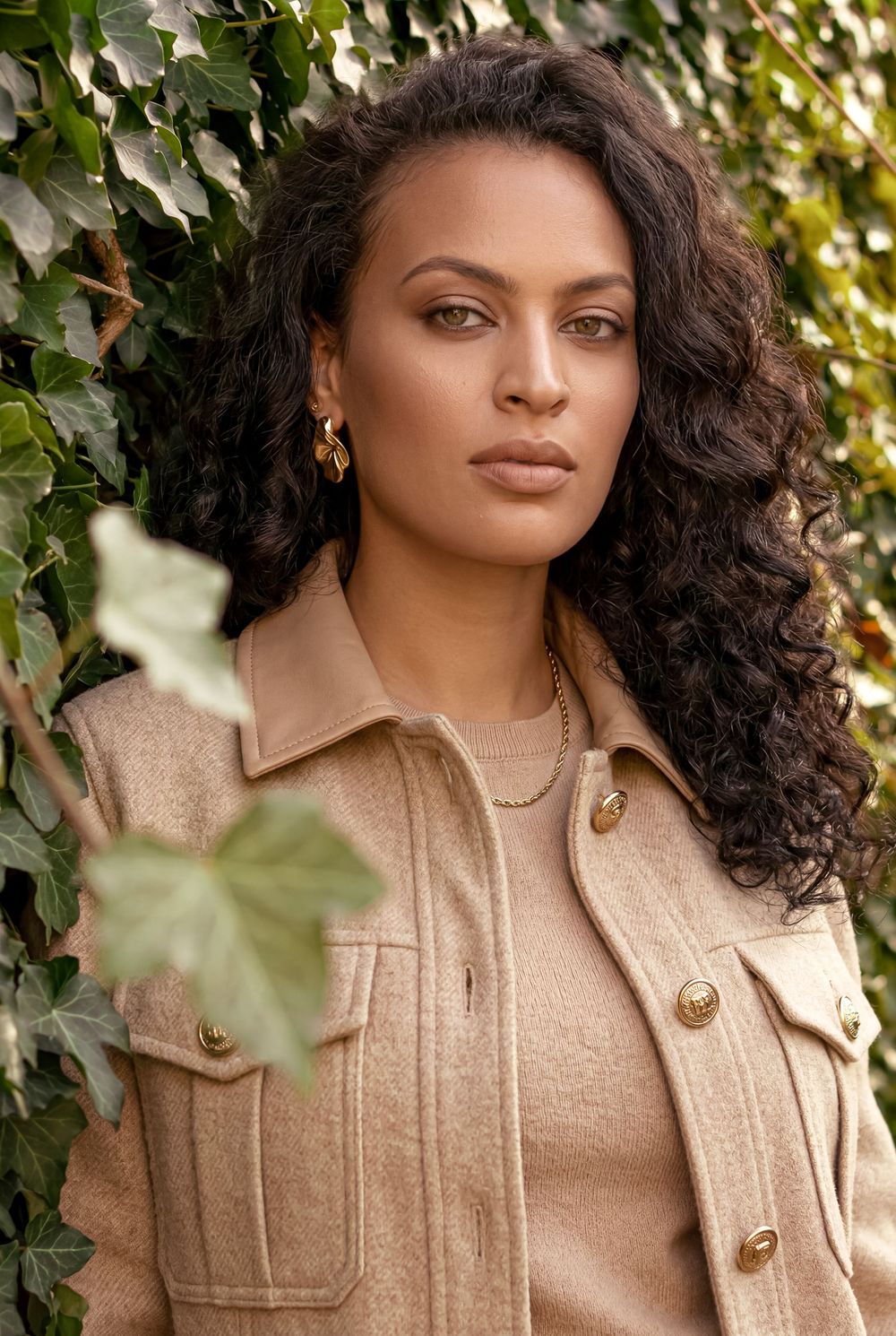 Woman with curly hair wearing a beige wool jacket and gold jewelry, posed against a green ivy wall.