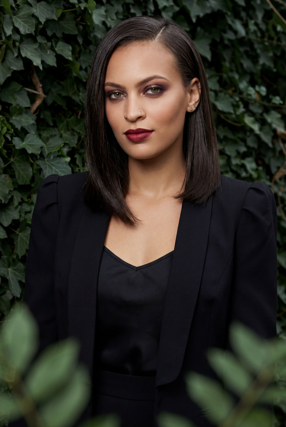 Woman in black blazer with dark hair and bold red lips poses against lush green ivy wall.
