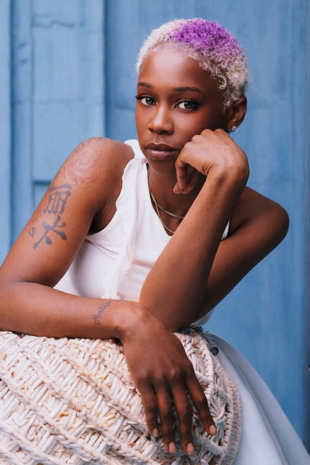 Portrait of someone with vibrant purple-tinted hair wearing a white tank top, leaning on a textured surface against a blue background.