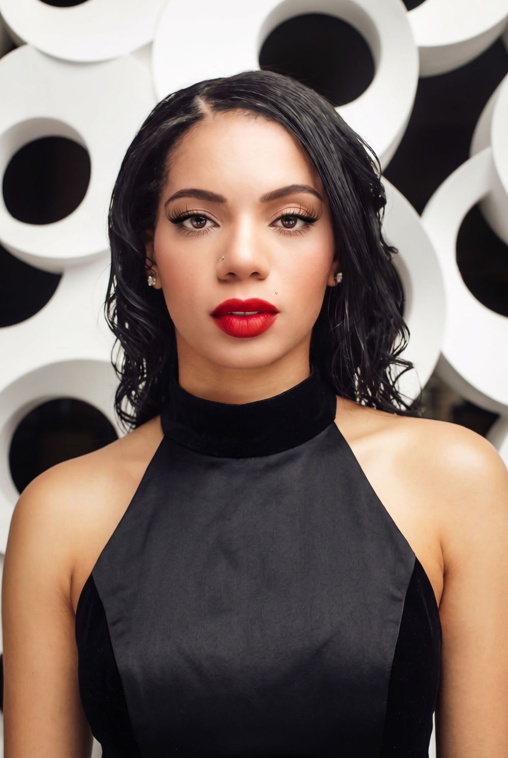 Young woman with red lips, black choker top, and dark curly hair poses against a white circular pattern backdrop.