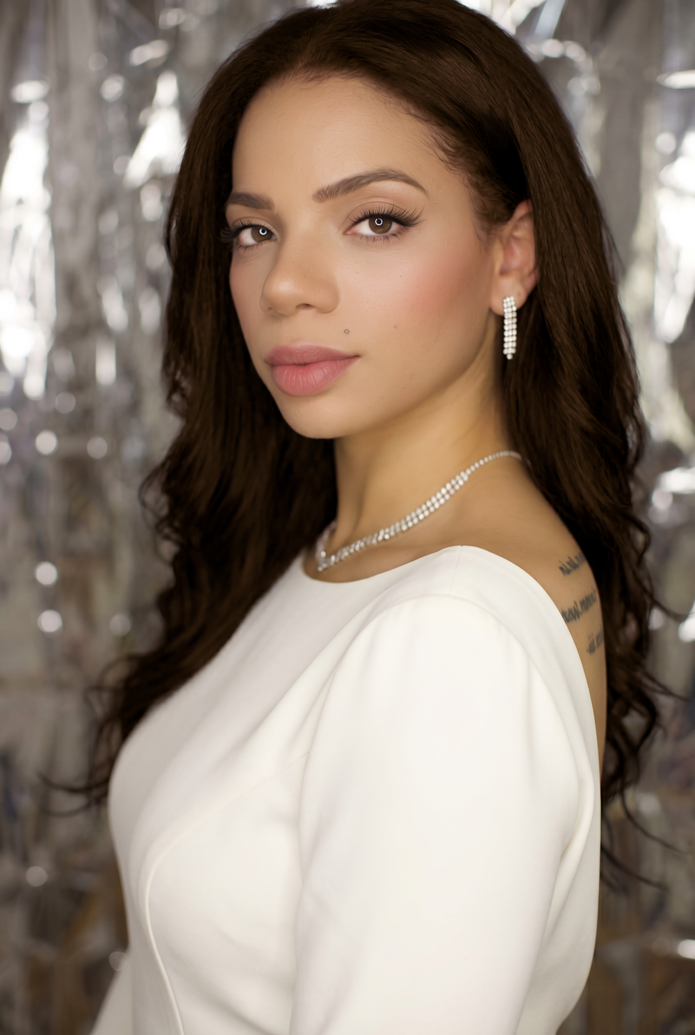 Young woman with long dark hair wearing a white dress, diamond earrings, and necklace posing against a silver backdrop.
