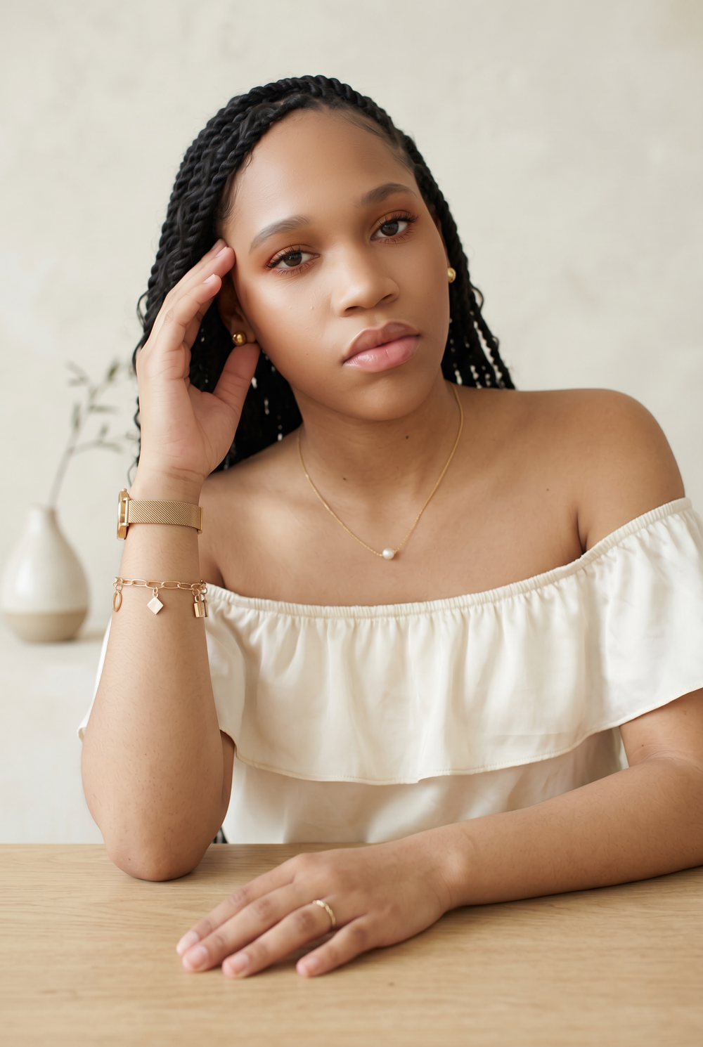 Young woman with braids wearing off-shoulder top and gold jewelry, posing thoughtfully at a wooden table.