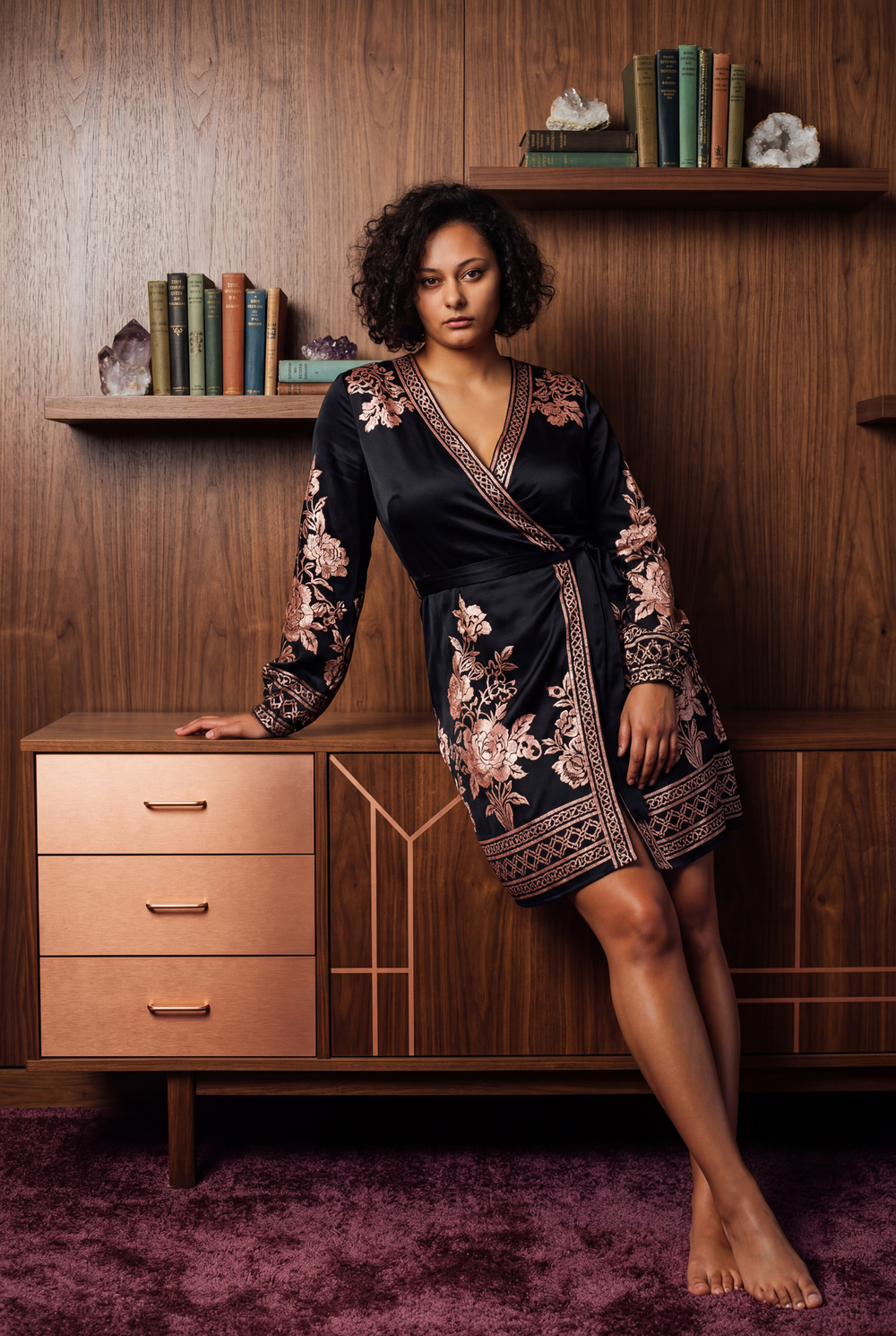 Woman in black floral embroidered wrap dress poses barefoot against mid-century modern wooden dresser with shelves.