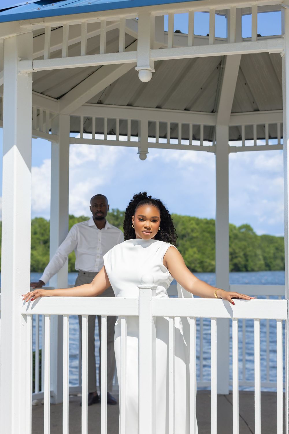 Woman in white dress posing in a gazebo by the water with a man standing in the background.