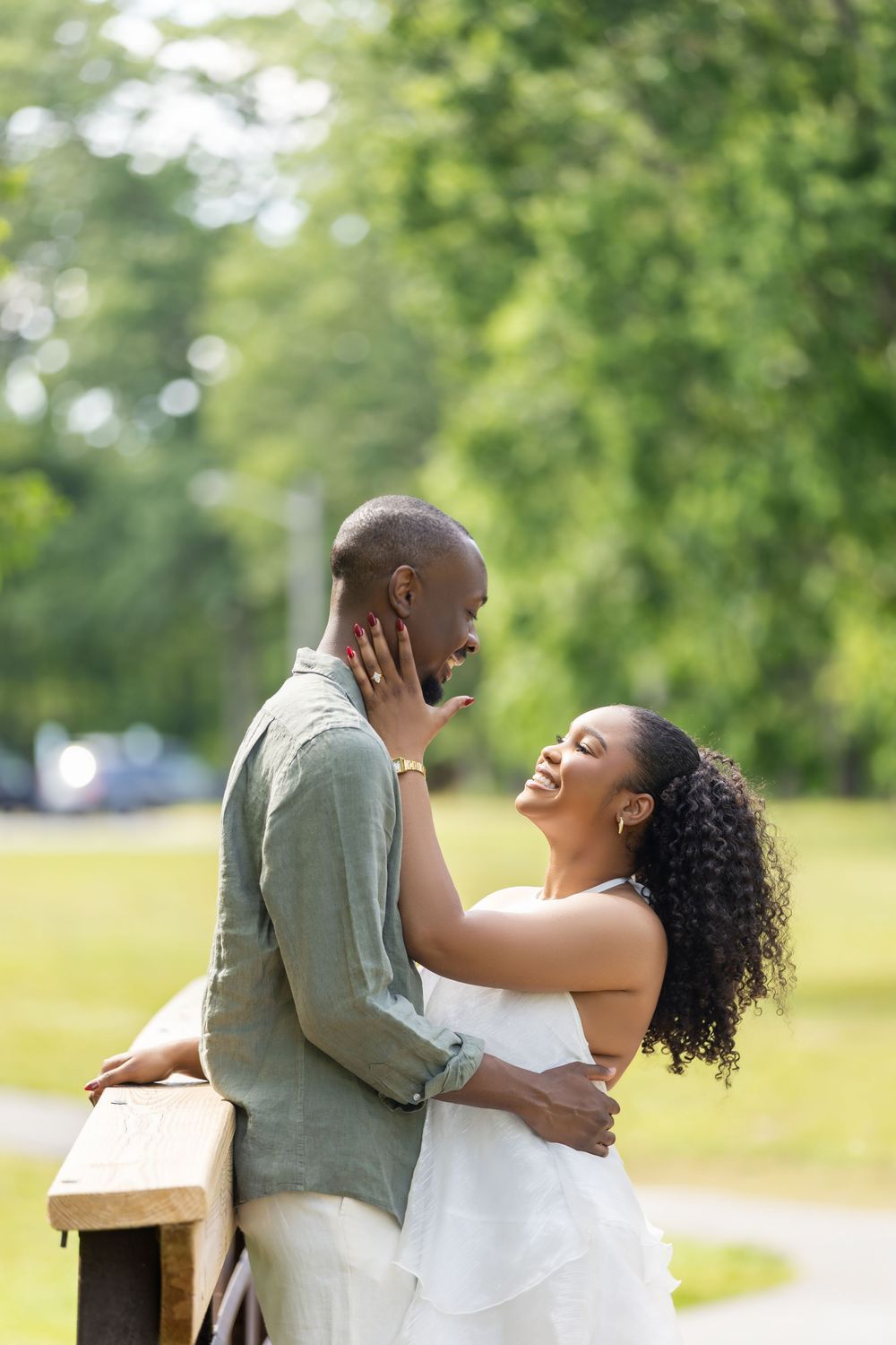 A joyful Black couple embraces in a sunny park, the woman in white laughing as she holds the man's face tenderly.