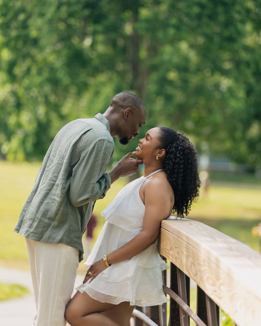Couple sharing a romantic moment on a wooden bridge surrounded by lush green trees in a park.
