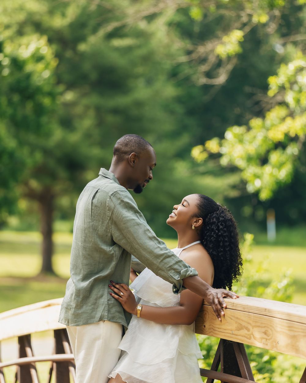 A couple shares a romantic moment on a wooden bridge surrounded by lush green trees in an outdoor engagement photo.