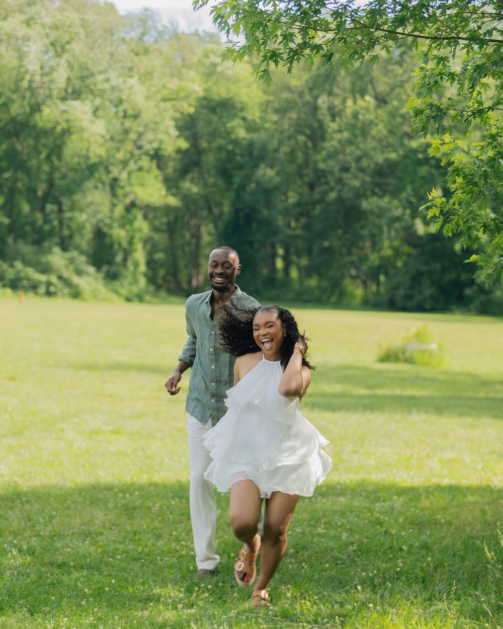 A joyful couple runs through a lush green field, the woman in a white dress laughing as the man follows behind her.