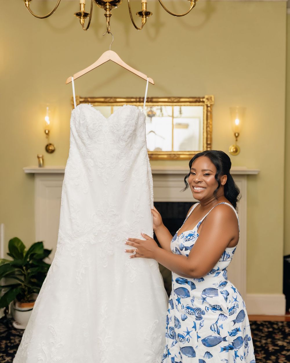 Smiling bride holding her white lace wedding gown on a hanger in an elegant room with chandelier.