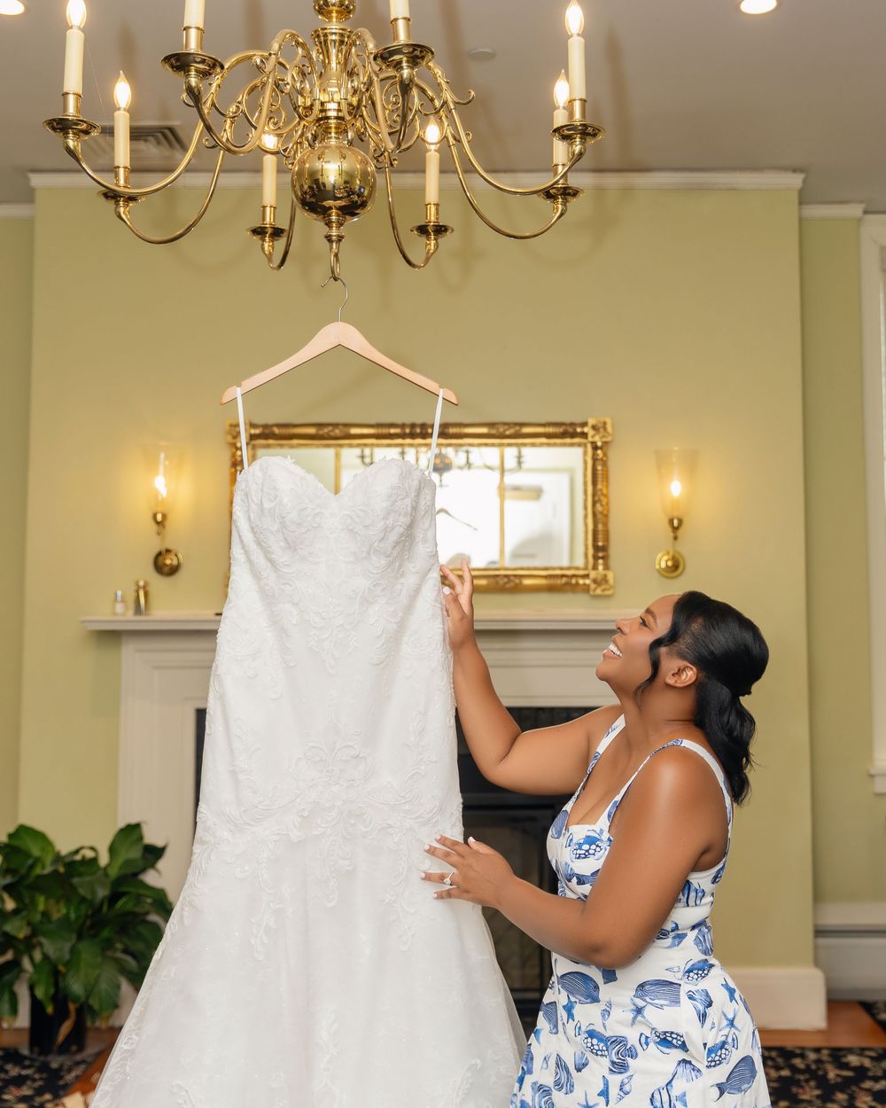 Bride admiring her white lace wedding gown hanging from a chandelier in an elegant room.