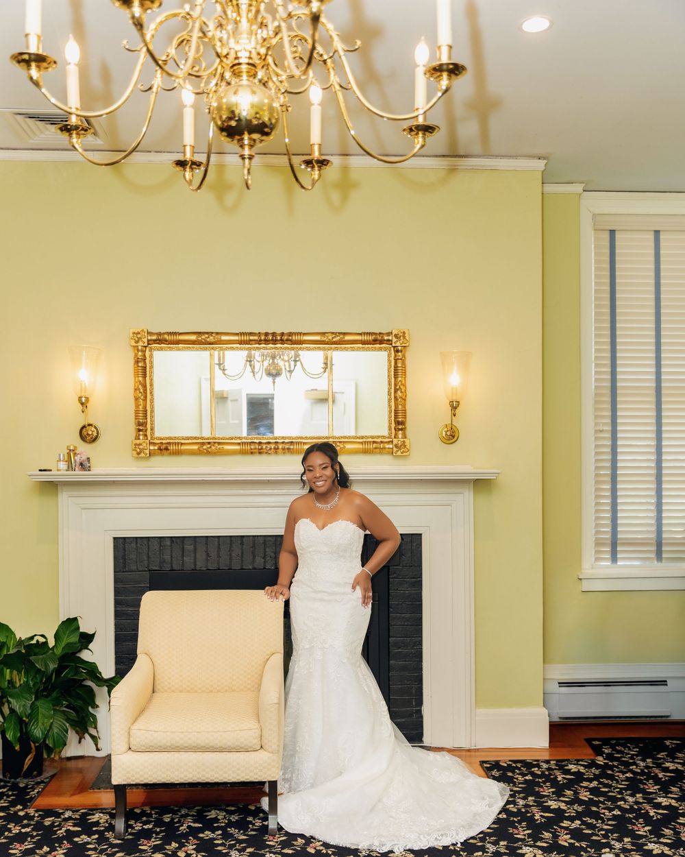 Bride in white mermaid gown poses by fireplace under gold chandelier in elegant yellow room.